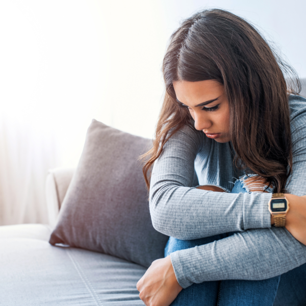 Young woman sitting on a sofa hugging her knees with her head bowed, looking sad