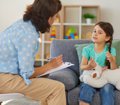 Therapist taking notes while a young girl with a teddy bear talks during a counseling session