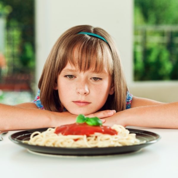 Child sitting at a table with a bored expression in front of a plate of pasta