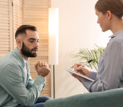 Man with clasped hands talking with a female therapist who takes notes during a counseling session